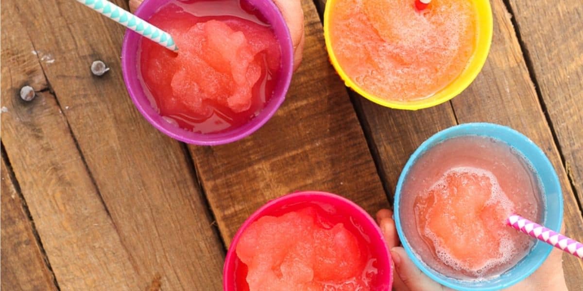 A top-view image of four colorful slushies in round cups on a wooden surface. The slushies are various shades of red and pink, and each cup has a striped straw in it. The cups are purple, yellow, blue, and pink. A hand is holding the blue cup at the bottom.