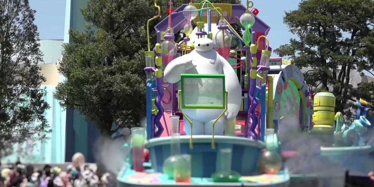 A parade float is decorated with a large figure of Baymax from Disney's "Big Hero 6." The float is vibrant and colorful, featuring various mechanical and scientific-themed elements. In the background, trees and a crowd of spectators are visible under a clear sky.