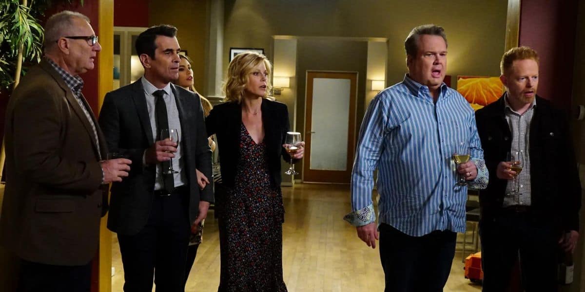 A group of people stands together in a room. They appear to be engaged in conversation, and some are holding drinks. The room has warm lighting, with a few doors and furniture visible in the background. The mood seems lively and social.
