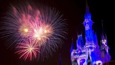 A nighttime scene at Tokyo Disney during Halloween features a beautifully illuminated castle with blue and purple lighting. Multicolored fireworks burst in the sky to the left of the castle, creating a magical and festive atmosphere.