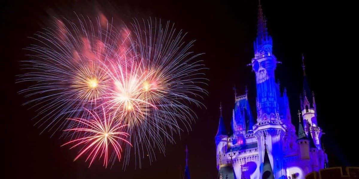 A nighttime scene at Tokyo Disney during Halloween features a beautifully illuminated castle with blue and purple lighting. Multicolored fireworks burst in the sky to the left of the castle, creating a magical and festive atmosphere.