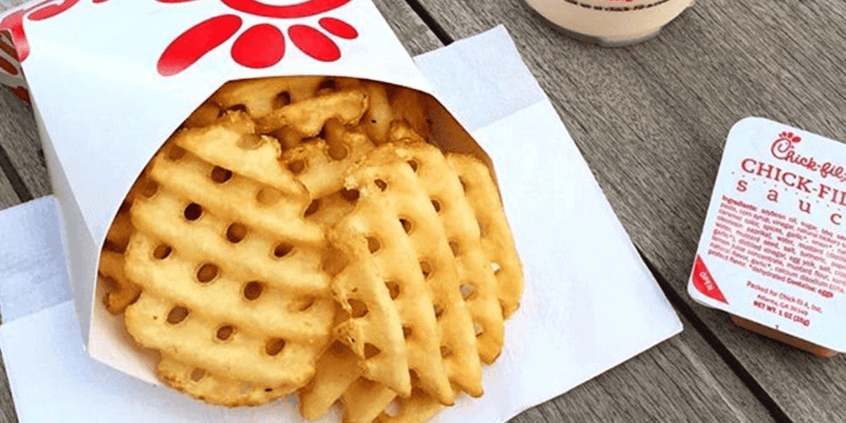 A serving of waffle fries is placed on a wooden table next to a container of Chick-fil-A sauce and a drink. The fries are in a white paper pouch with the Chick-fil-A logo. Enjoy them while you can, as there are rumors about Chick-fil-A getting rid of waffle fries soon!