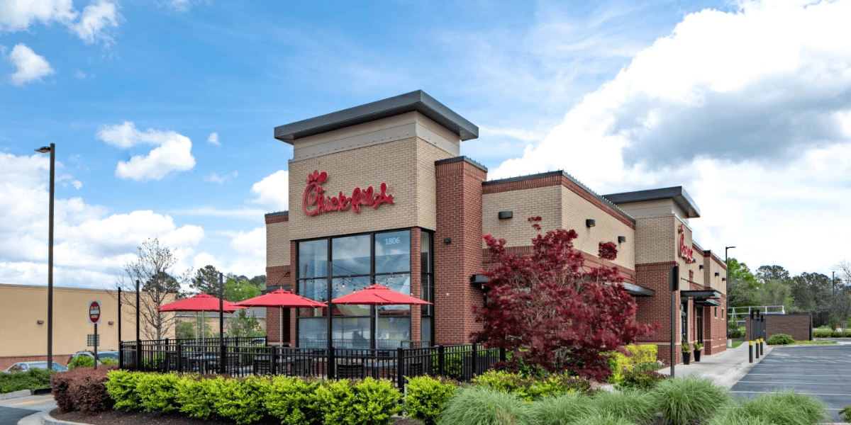 A Chick-fil-A restaurant features a modern brick exterior, black awnings, and red umbrellas on the outdoor seating area. The entrance has the signature Chick-fil-A logo, despite rumors of Chick-fil-A getting rid of waffle fries. Surrounding the restaurant are neatly manicured shrubs and trees under a partly cloudy sky.