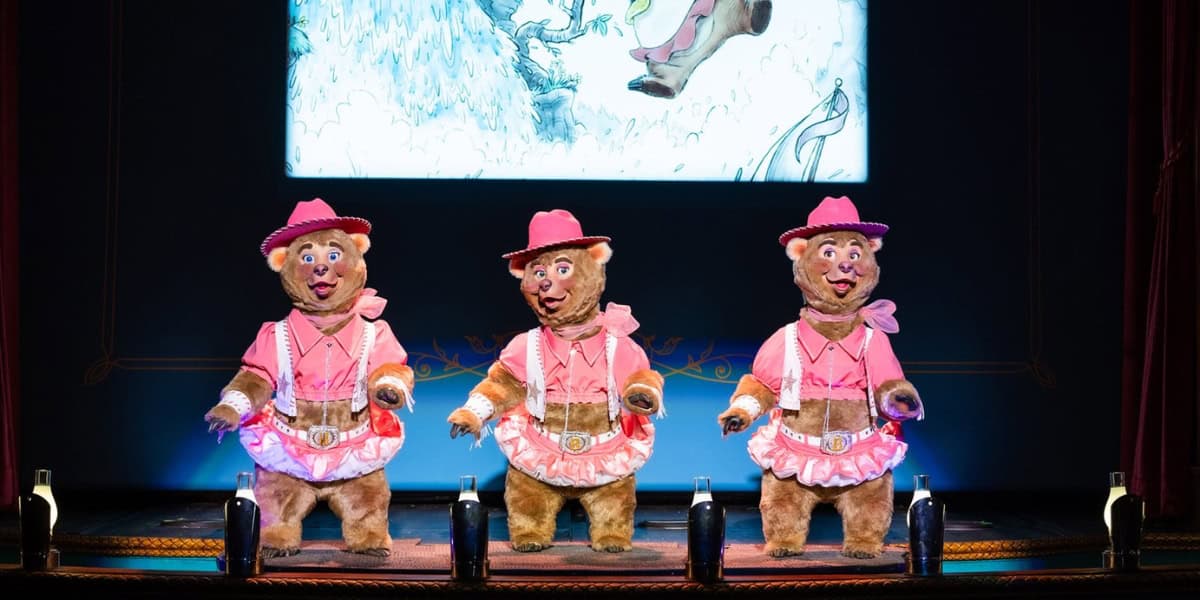 Three bear characters, dressed in pink hats and western outfits, are performing on a stage. They are standing side by side, with a backdrop featuring a cartoon drawing. The stage is framed with red curtains, and there are spotlights illuminating the scene.