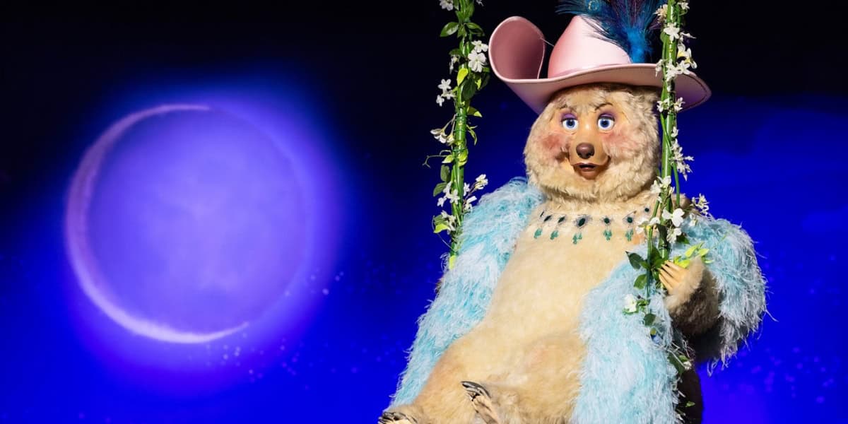 A bear puppet wearing a pink hat with feathers, blue necklace, and matching blue fur coat sits on a swing decorated with flowers, against a backdrop of a glowing purple moon.