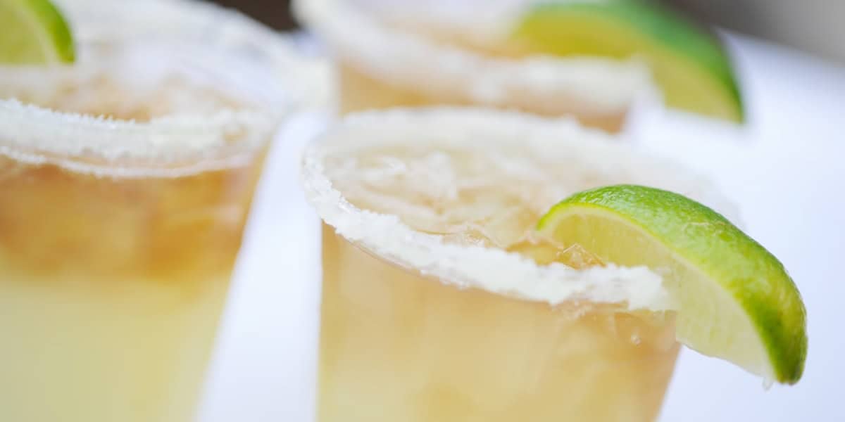 Close-up of three margaritas with salted rims. The glasses are filled with a light, icy beverage, and each has a lime wedge garnish on the rim. The background is out of focus, highlighting the drinks in the foreground.