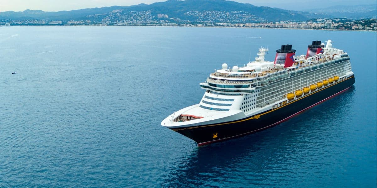An expansive cruise ship, the Disney Dream, sails through calm blue waters with a hilly coastline and a city in the distance under a cloudy sky. The ship features multiple decks and lifeboats, painted in navy blue, white, and red.