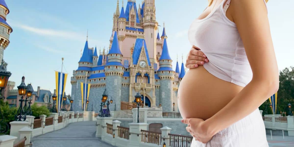 A pregnant woman in a white outfit stands holding her belly in front of Cinderella's Castle at a Disney theme park.