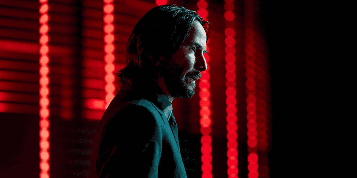 A person with shoulder-length dark hair and a beard is shown in profile against a backdrop of vertical red illuminated lines. The individual is wearing a dark jacket, and the scene has a dramatic and intense lighting effect.