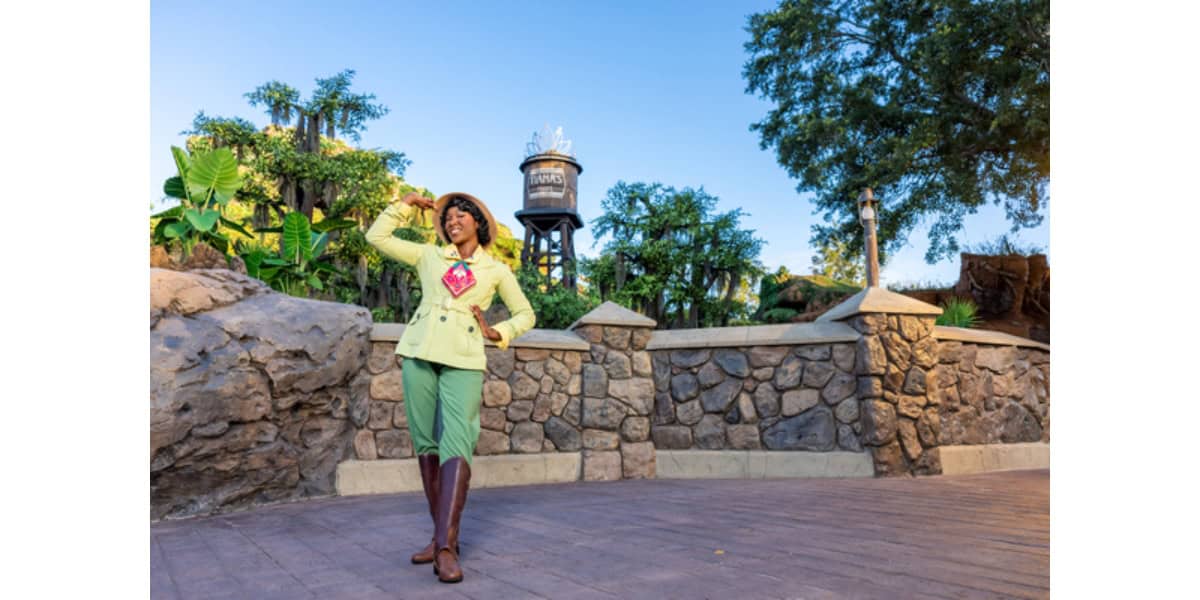 A Disney character performer in a yellow jacket and green pants, wearing brown boots and a brown hat, stands on a paved path. They are striking a pose with a raised hand near their head. Behind them is a stone wall, lush greenery, and a tall water tower. The sky is clear and blue.