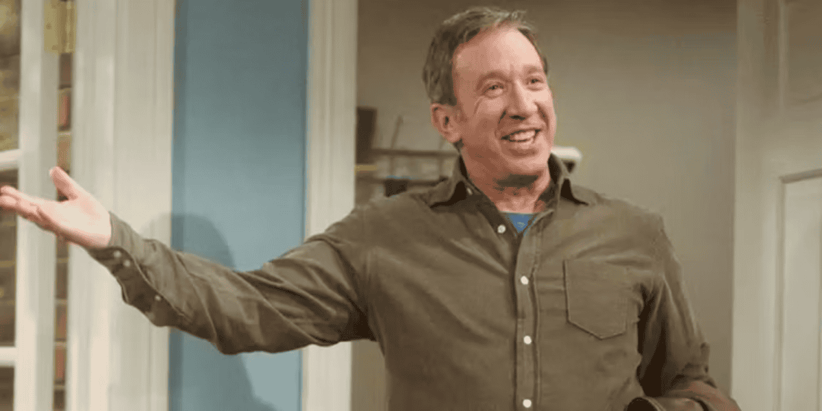 A man with short hair is standing indoors, smiling broadly and holding a leather jacket in his left hand. He is wearing a brown button-up shirt over a blue undershirt. His right arm is extended in a welcoming gesture. The background includes a partially visible shelf.