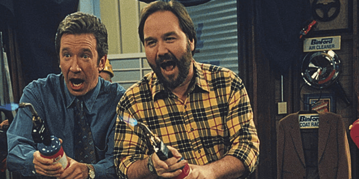 Two men are holding flamethrowers and smiling excitedly. The man on the left is wearing a blue shirt and tie, while the man on the right is wearing a yellow plaid shirt. They are in a workshop setting with tools and signs in the background.