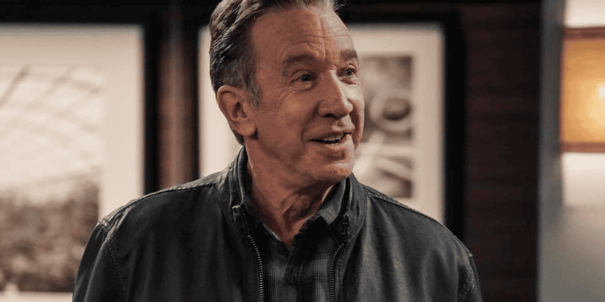 A man with short, graying hair and a light smile is wearing a dark leather jacket over a blue plaid shirt. He is indoors, standing in front of a wall with framed black-and-white photographs. The lighting is warm and gives a cozy ambiance to the scene.