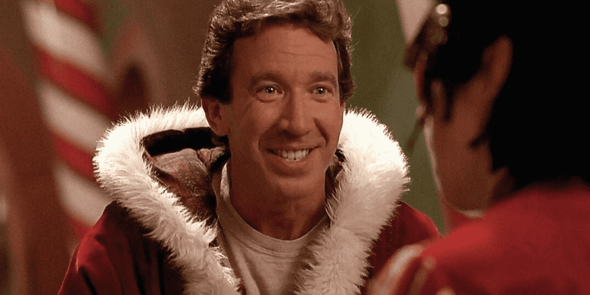 A smiling person dressed as Santa Claus with a red suit and white fur trim sits indoors. Blurred background with festive décor hinting at a Christmas setting. The individual is facing another person who is partially visible from behind.