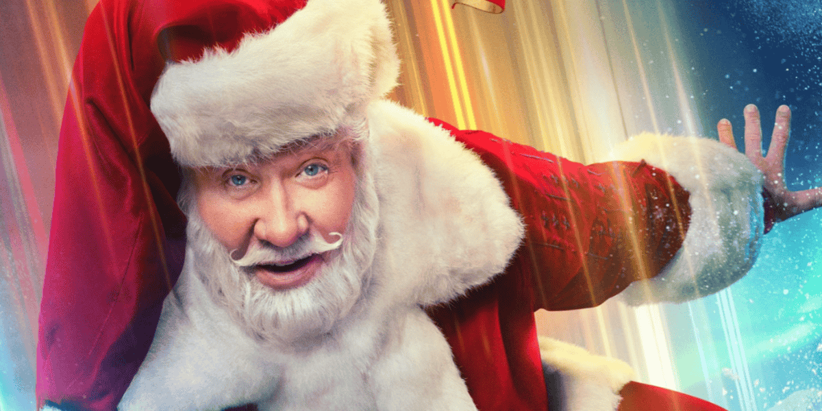 A person dressed as Santa Claus, with a fluffy white beard and mustache, smiles warmly. He is wearing a traditional red and white Santa suit with a matching hat, striking a playful pose with one arm extended. The background features vibrant, colorful lights.