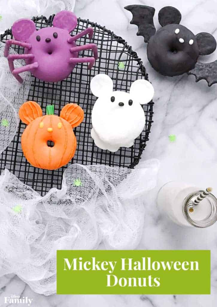 Four Halloween-themed donuts are arranged on a black cooling rack. The donuts resemble a spider, bat, pumpkin, and a ghost, each decorated with playful features. A bottle of milk with a striped straw is nearby, set on a marble surface.