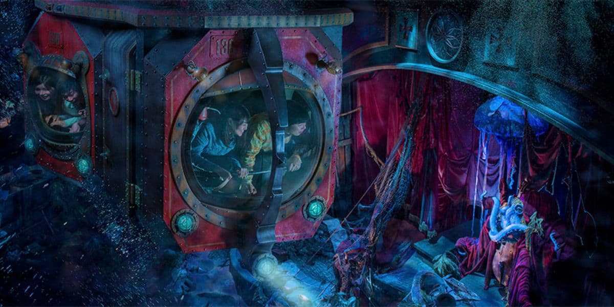 A vibrant, underwater-themed amusement park ride with two submarine pods filled with excited riders. The scene features red curtains, colorful lights, aquatic decorations, and an animatronic figure dressed in vibrant costume amidst the water-themed background.