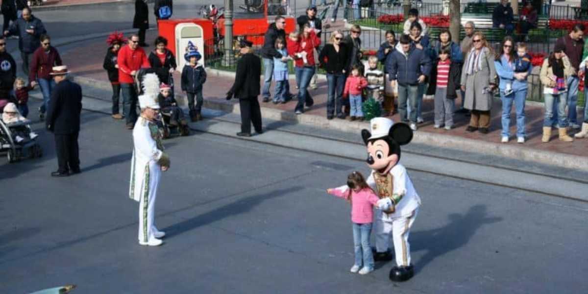 A parade scene at a theme park shows a crowd of adults and children watching. A performer dressed as Mickey Mouse and a marching band leader interact with a young girl who is pointing at the ground. The parade is taking place in a festive, lively environment.