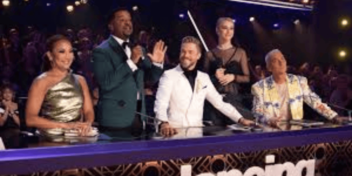 Five judges are standing behind a panel table on a brightly lit stage. From left to right: a woman in a metallic gold dress, a man in a teal suit holding a microphone, a man in a white suit, a woman in a black dress, and a man in a colorful jacket. An audience is visible in the background.
