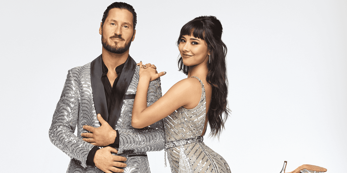 A man and a woman pose together against a plain white background. The man wears a sparkly silver jacket with a black shirt, while the woman is dressed in a glittery silver dress. Both look directly at the camera, with the woman resting her hand on the man's shoulder.