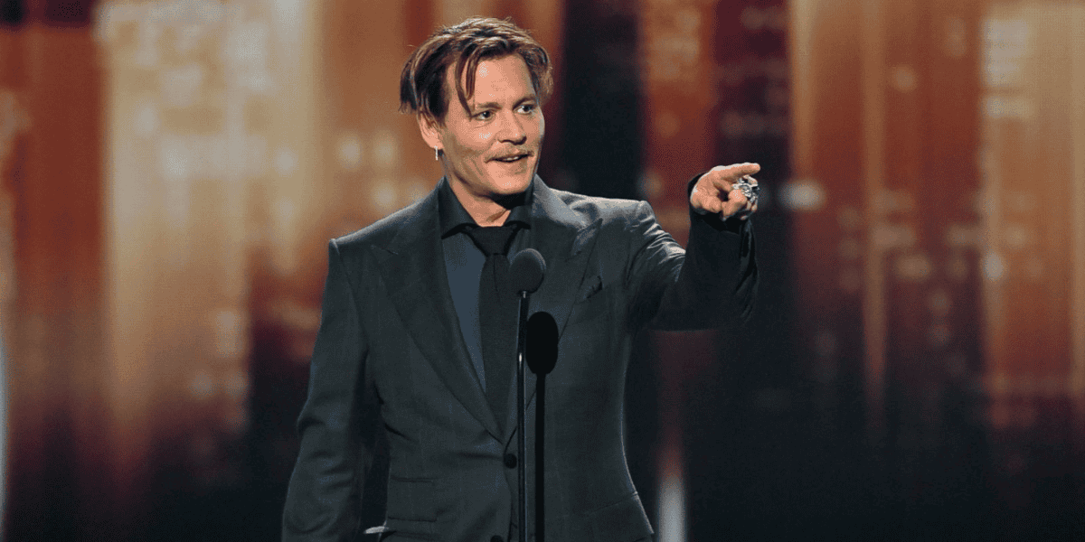 A person with medium-length hair stands on a stage, wearing a dark suit and a dark shirt underneath. They are gesturing with their right hand while speaking into a microphone about Johnny Depp's new role. The background is a blurred mix of warm colors, giving a shimmering effect.