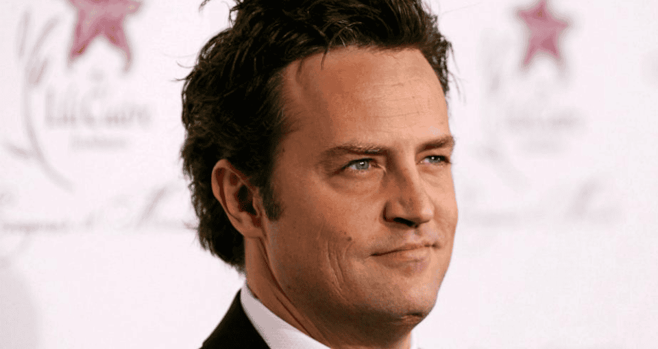 A man with short, dark, tousled hair is pictured in close-up. He is wearing a dark suit jacket, white shirt, and a subtle smile with a neutral expression. The background is light-colored with indistinguishable text and a red star logo.
