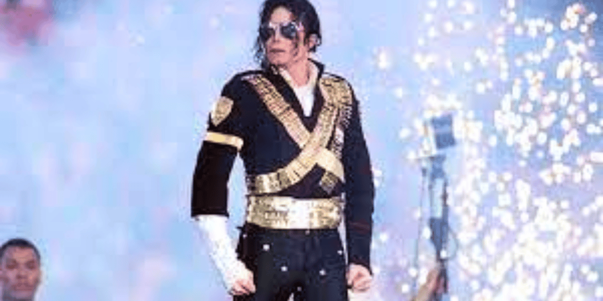 A performer stands on stage wearing a black jacket adorned with gold details, sunglasses, and a single white glove. The background features bright, colorful lighting and fireworks, emphasizing a dynamic and energetic atmosphere.
