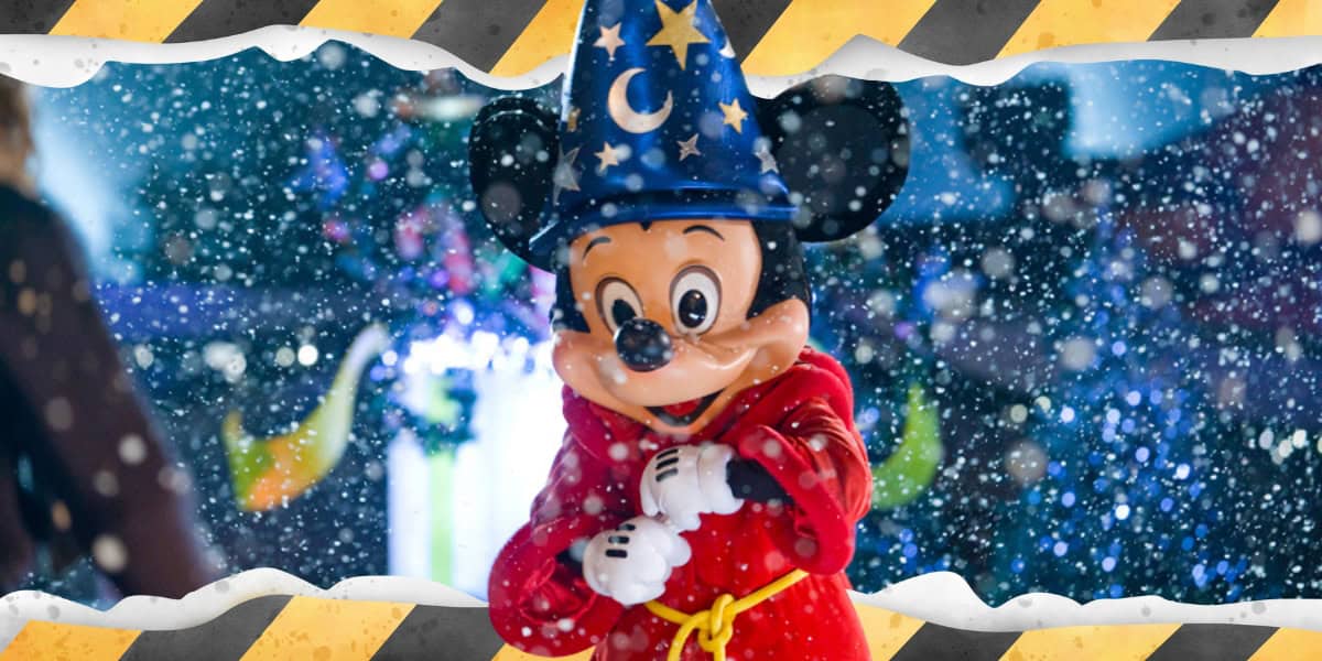 Mickey Mouse wears a blue wizard hat and red robe with a yellow tie while standing in a snowy scene. The background features blurry lights and decorations. The image is bordered by a yellow and black striped frame.