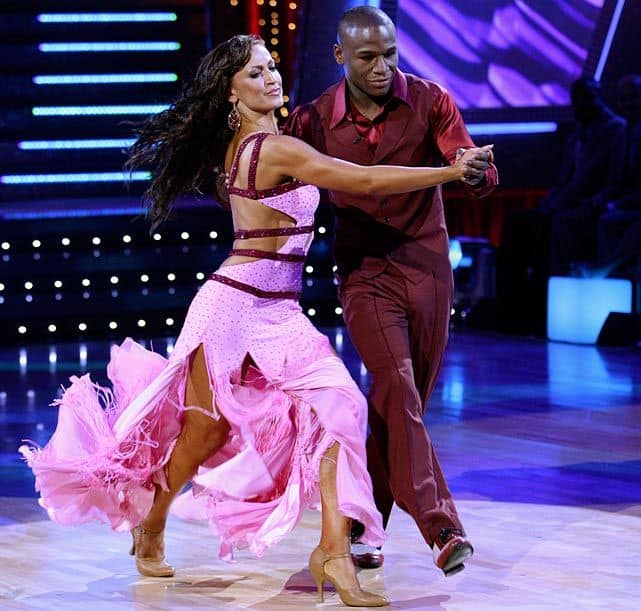 A man and a woman are dancing energetically on a lit dance floor, reminiscent of Dancing with the Stars. The woman wears a flowing pink dress with cutouts, and the man wears a maroon shirt and pants. They are mid-step, with the woman's dress moving gracefully and lights in the background.