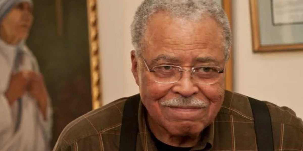 An older man with short, curly gray hair, mustache, and glasses smiles slightly while wearing a brown plaid shirt with black suspenders. In the background, there is a painting of a woman in white attire with a blue border.
