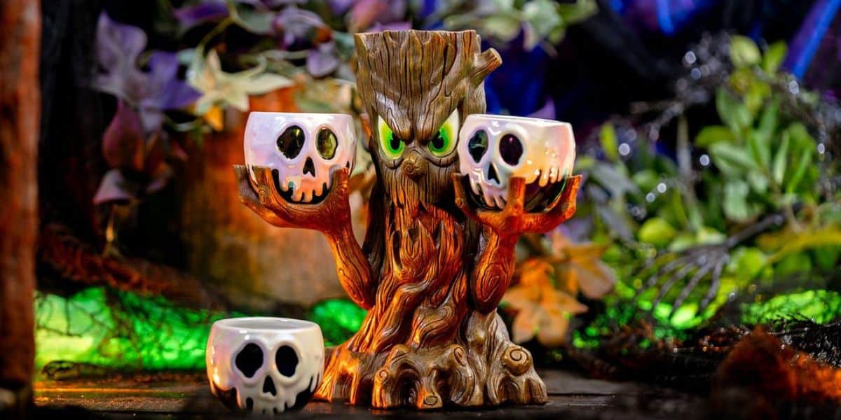 A spooky, tree-like figurine with an angry face and glowing green eyes holds two skull-shaped mugs in its branches. Another skull-shaped mug sits nearby. The background features eerie green and blue lighting with tangled vines and leaves.