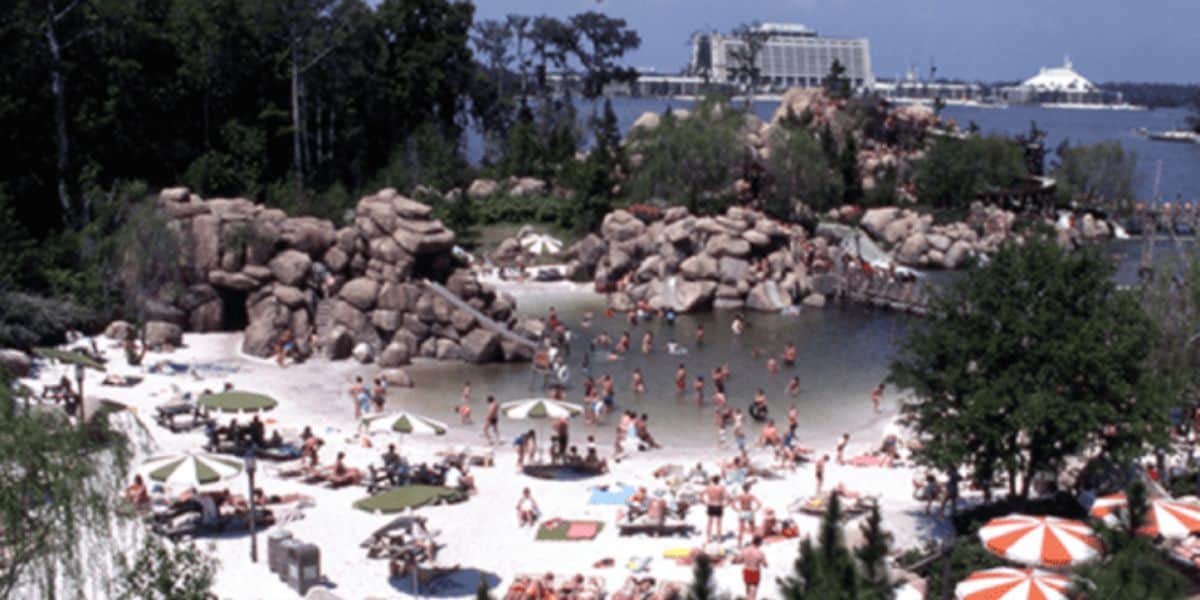 A busy beach scene with numerous people sunbathing, swimming, and lounging under umbrellas on white sand. Rocky formations are scattered around the beach area. A body of water and a large building are visible in the background. Trees and greenery surround the area.