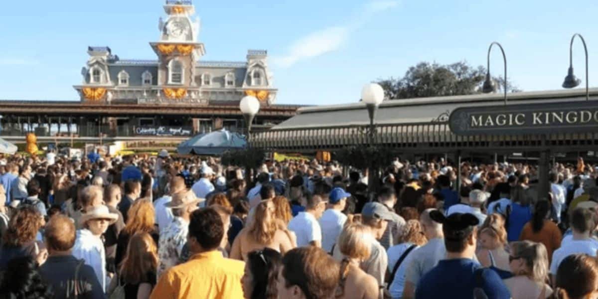 A large crowd of people gather in front of the entrance to Magic Kingdom, with the Main Street train station visible in the background. The entrance area is bustling with visitors dressed in various outfits, some wearing hats, and a clear sky overhead.