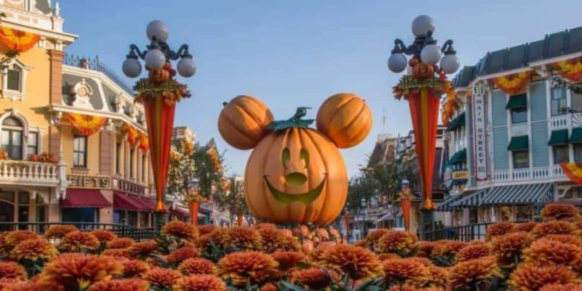 A giant Mickey Mouse-shaped pumpkin with a green leaf on top is the centerpiece of a festive plaza decorated for Halloween. Orange flowers cover the foreground, and themed lampposts adorned with orange garlands frame the scene. Victorian-style buildings line the street.