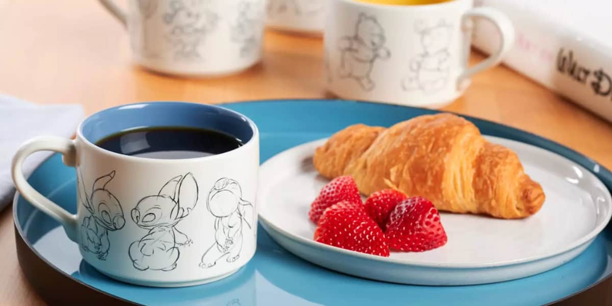 A breakfast setting features a blue tray holding a white cup filled with coffee. The cup has sketches of Disney characters on it. Beside the cup is a plate with a croissant and several fresh strawberries. Additional mugs with sketches and an open book are in the background.