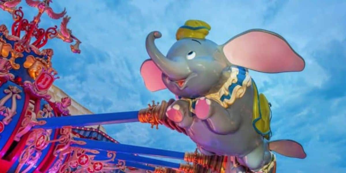 A colorful amusement park ride in the heart of Disney World features a large, smiling, flying elephant with big pink ears and a blue and yellow cap. Amidst lively Disney World crowds, the ride's ornate details shine beautifully against a blue sky dotted with clouds.
