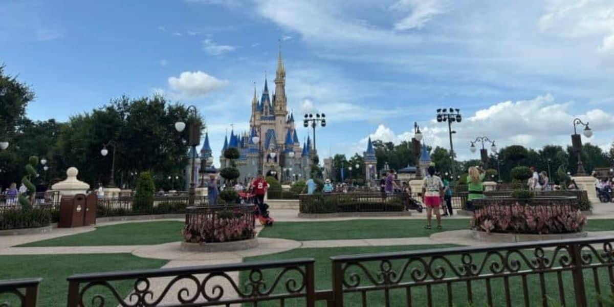 A wide view of a theme park shows Cinderella Castle in the background. People are walking and relaxing on a large green lawn with flower beds and ornate fencing. The sky is partly cloudy, enhancing the picturesque scenery.
