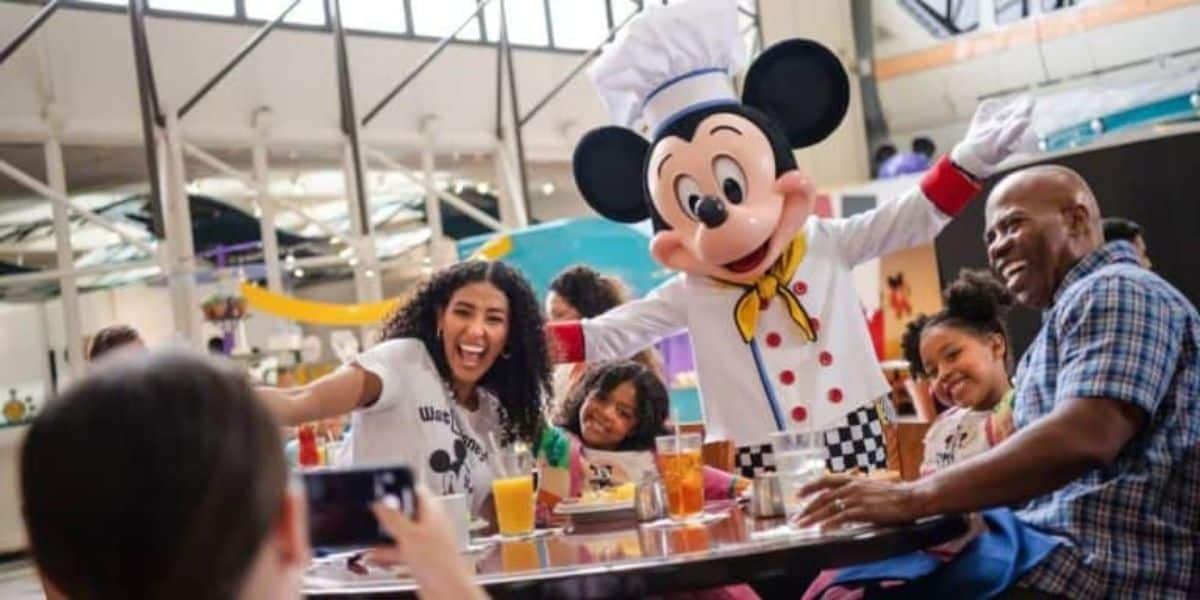A family with two young children enjoys a meal at a restaurant, posing for a photo with a person dressed as a chef Mickey Mouse. They are all smiling and appear to be having a great time. The setting looks lively and cheerful with large windows in the background.