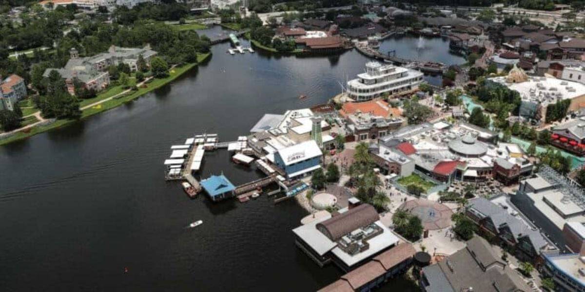 Aerial view of a bustling waterfront town featuring various buildings, docks, and a prominent riverboat. The area includes shops, restaurants, boats docked at piers, and surrounding green spaces, all adjacent to a large body of water.