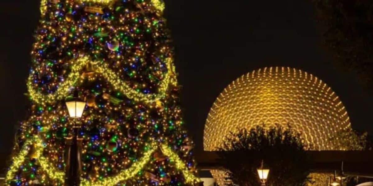 A large Christmas tree adorned with colorful lights and ornaments stands illuminated at night. Behind it, a geodesic dome structure glows with warm yellow light. Street lamps and trees frame the festive scene.