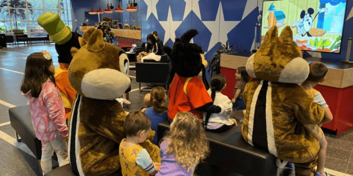 A group of children sit on benches in a bright, star-themed room, watching an animated cartoon on a large screen. They are accompanied by people wearing costumes of famous animated characters, including a dog, a chipmunk, and other animal figures.