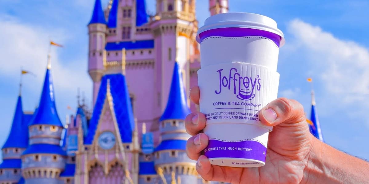 A hand holds a Joffrey's Coffee & Tea cup in front of a brightly colored castle with blue and pink turrets under a clear sky. The sleeve on the cup reads, "Joffrey's Coffee & Tea Company".
