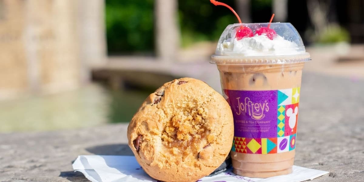 A large chocolate chip cookie resting on a white paper wrapper next to a clear plastic cup of iced coffee topped with whipped cream and cherries. The cup has a colorful label with the Joffrey's Coffee & Tea Company logo. Both items are on a stone surface.