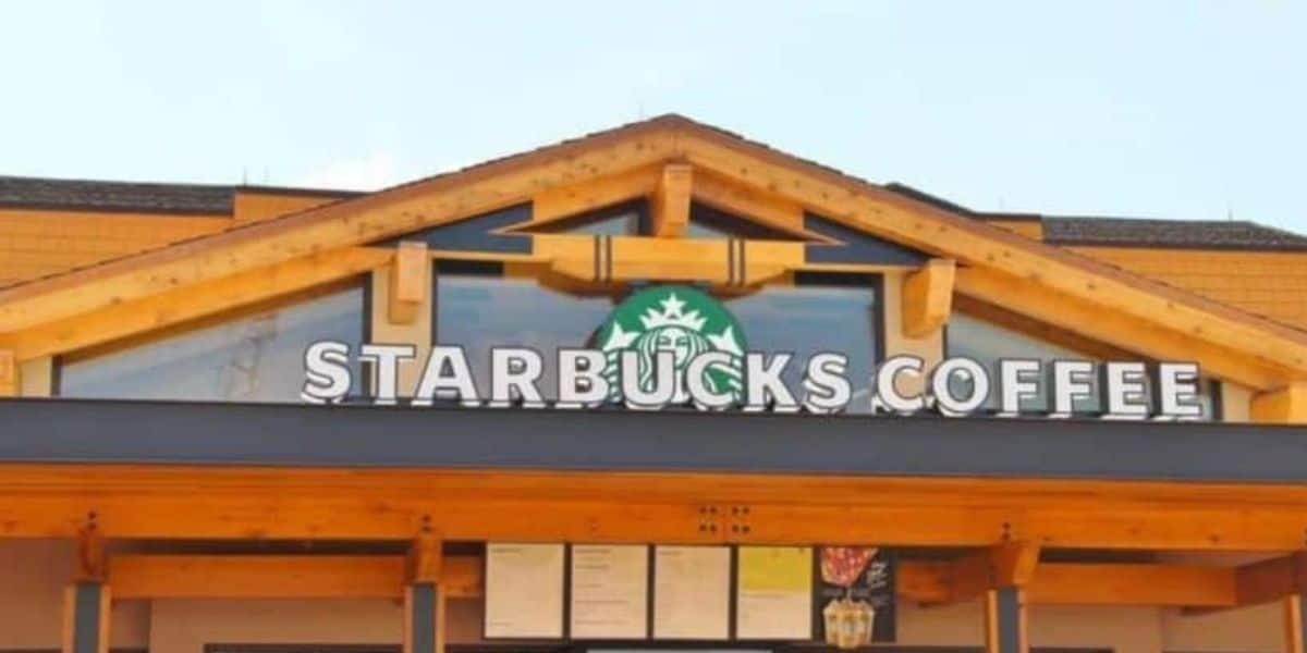 A storefront of a Starbucks Coffee shop features a large green Starbucks logo and illuminated capital letters spelling "STARBUCKS COFFEE." The building has a wooden exterior with a peaked roof, and the sky is clear in the background at Disney World.