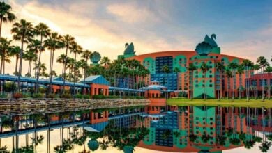 A large resort building styled with whimsical architectural elements reflecting in a calm body of water. The building has green swan sculptures on the roof and is surrounded by tall palm trees. The sky is colorful with hues of blue, orange, and yellow during sunset at Disney World.