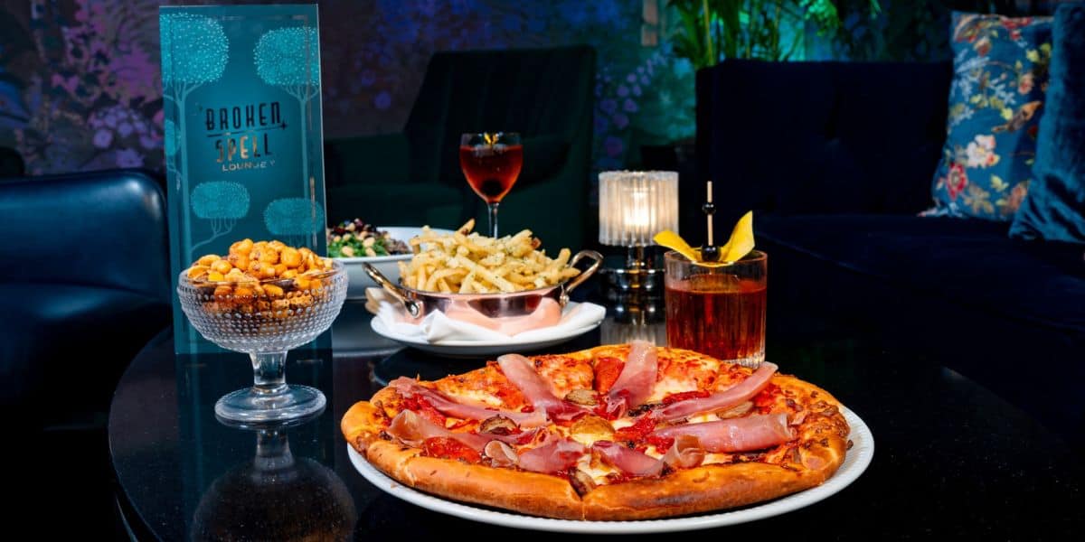A variety of dishes on a table include a pizza topped with ham, a bowl of mixed nuts, a plate of seasoned fries, and a salad. Two cocktails are in the background on a dimly lit table with a Broken Seal Lounge menu.