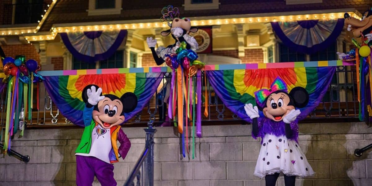 Mickey and Minnie Mouse, dressed in colorful costumes, wave alongside Goofy. They are in front of a decorated building with rainbow banners and lights, celebrating a festive event.