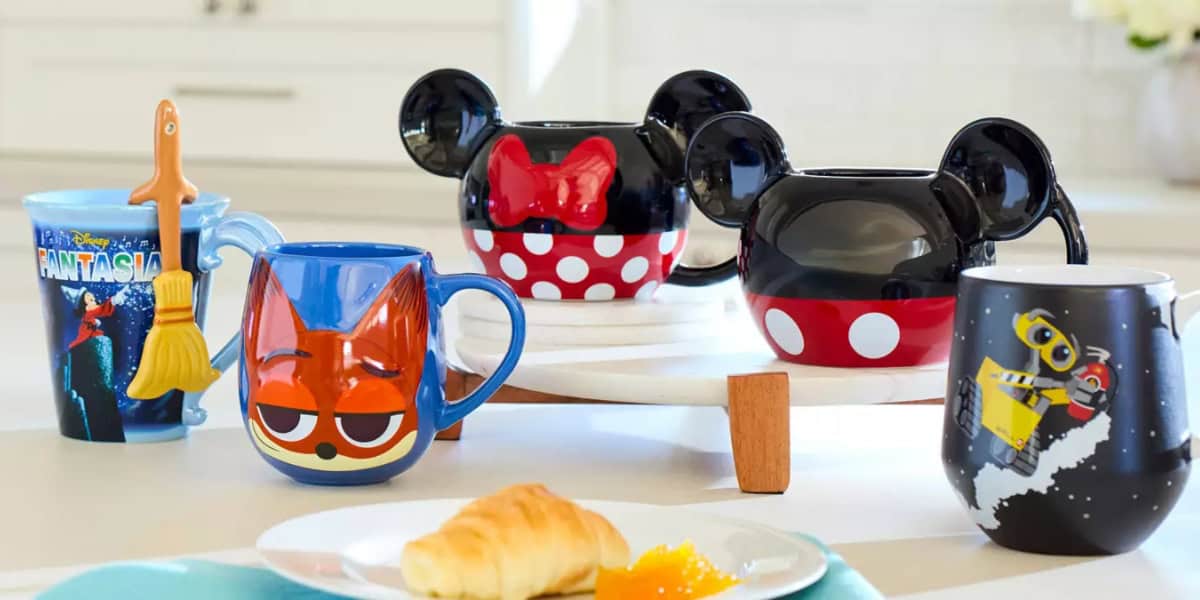 A collection of colorful Disney-themed mugs on a table, featuring designs from Fantasia, Zootopia, Mickey and Minnie Mouse, and WALL-E. In the foreground, there's a plate with a croissant and marmalade.