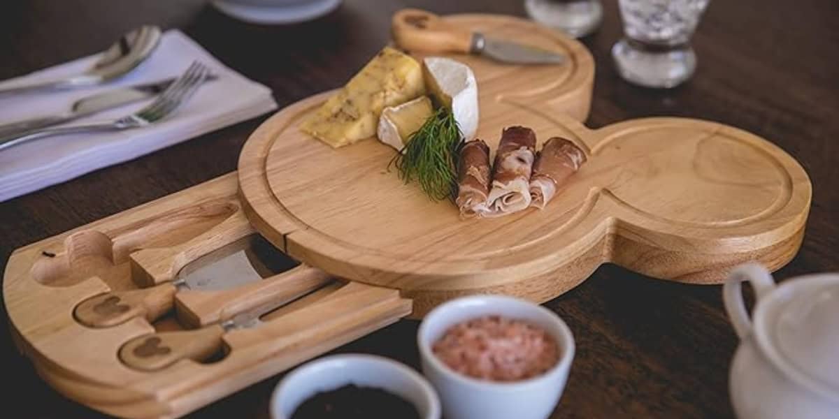 A wooden, Mickey Mouse-shaped cheese board with various cheeses and rolled meats placed on top. The board features compartments containing cheese knives. Surrounding it are utensils, a teapot, and small bowls containing different condiments.