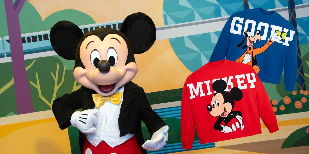 Mickey Mouse poses in front of a colorful backdrop featuring a monorail and geometric shapes. Next to him, two sweatshirts are displayed: one blue with Goofy and the word "GOOF," and one red with Mickey and his name written above.